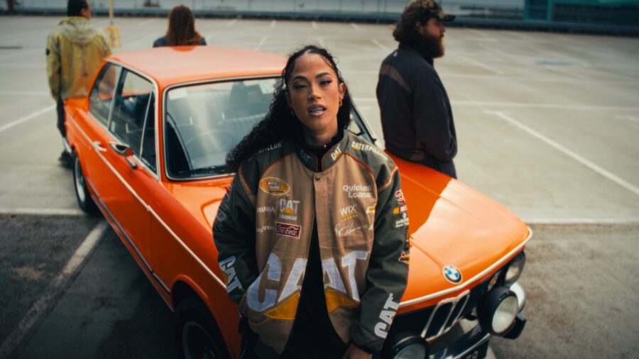 A singer in a motorsport jacket leaning against an orange car in an empty car park