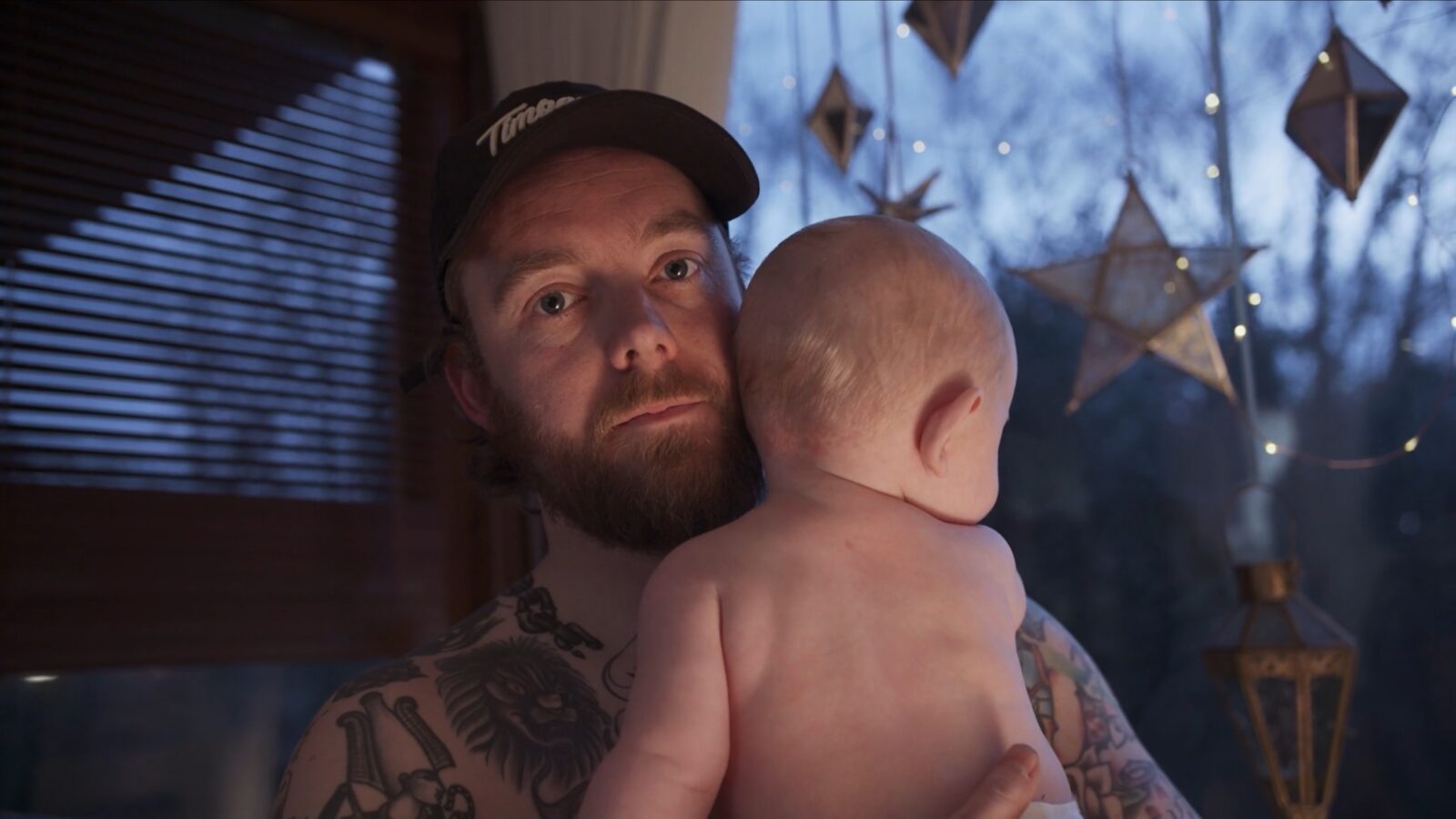 A tattooed man holding his baby at a window hung with paper stars