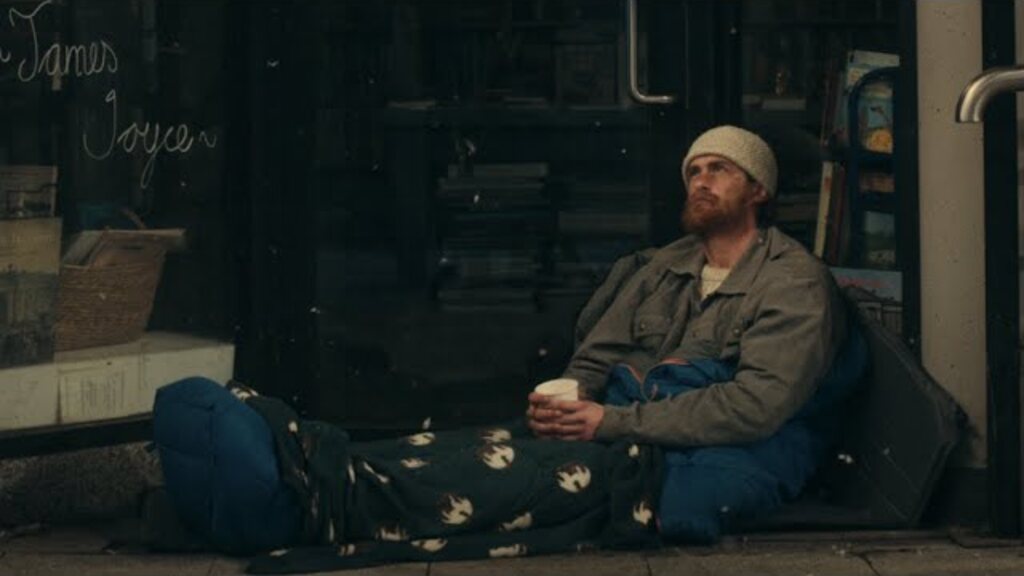 A man in a beanie sitting on the pavement outside a shuttered Dublin shopfront with a cup in his hands