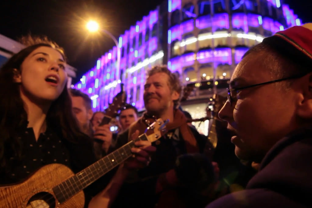 Musicians singing together at night, a building lit purple behind them