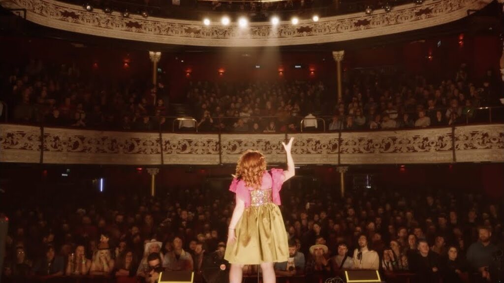 CMAT alone on a theatre stage with an arm raised, the tiered balconies packed behind her