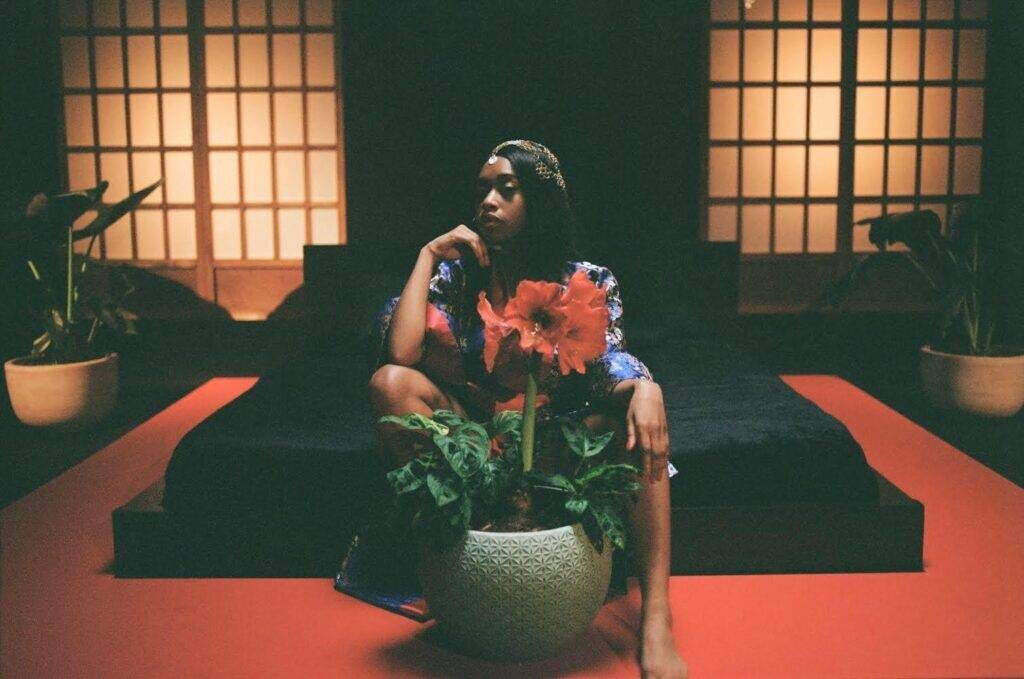 Denise Chaila seated beside a large orange flower on a red-lit platform, low light through paper screens behind her