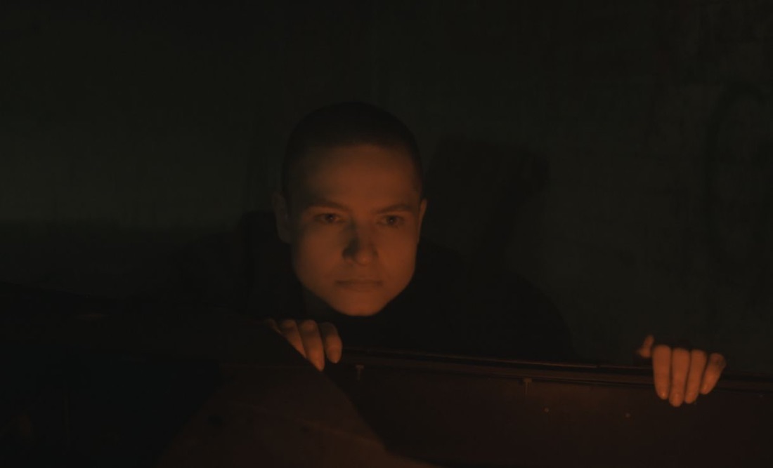 A face lit by orange light, hands gripping a wooden ledge in the dark