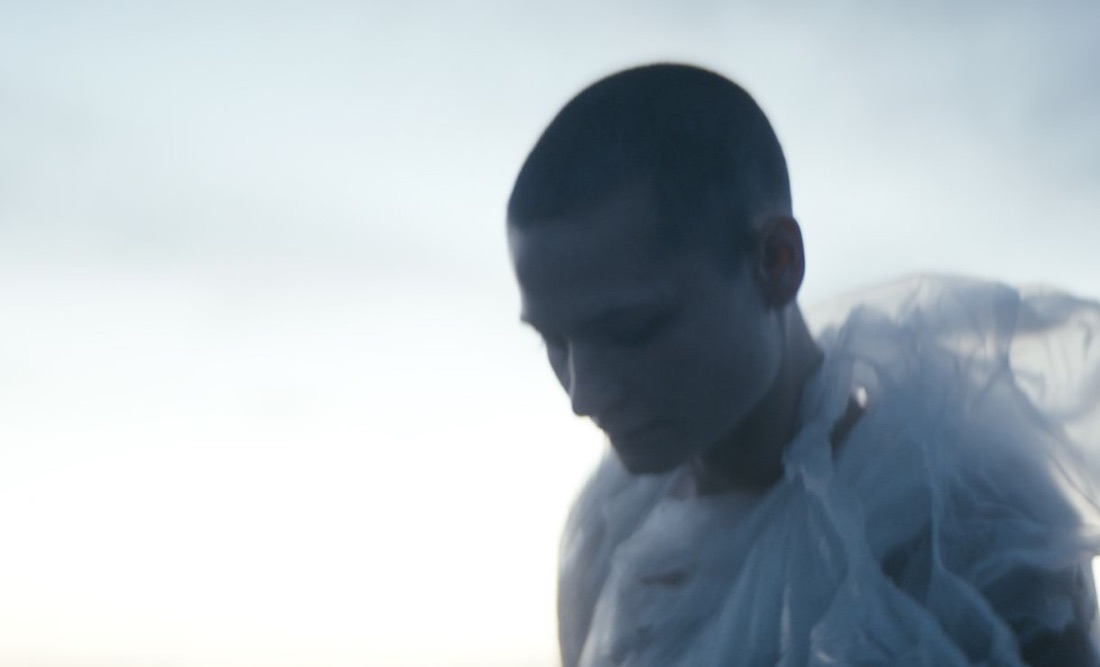 A figure wrapped in translucent plastic sheeting against a pale sky