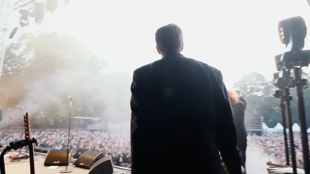 A musician seen from behind on stage, facing a packed festival crowd through hazy evening light