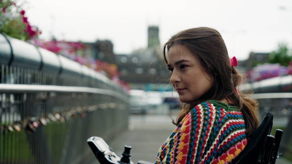 A woman in a wheelchair looking out over a Dublin bridge decorated with padlocks, city buildings behind her