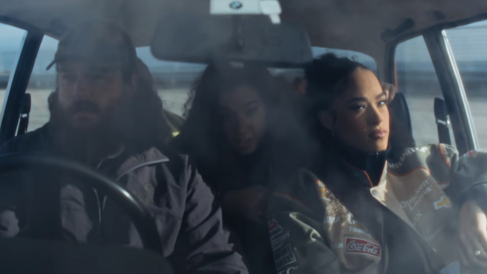 A singer in a motorsport jacket leaning against a red car in an empty rooftop car park, the words Can't Deny It across the frame
