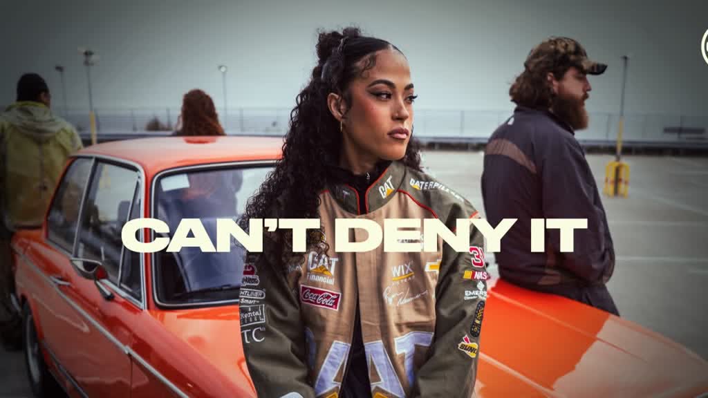 A singer in a motorsport jacket leaning against a red car in an empty rooftop car park, the words Can't Deny It across the frame