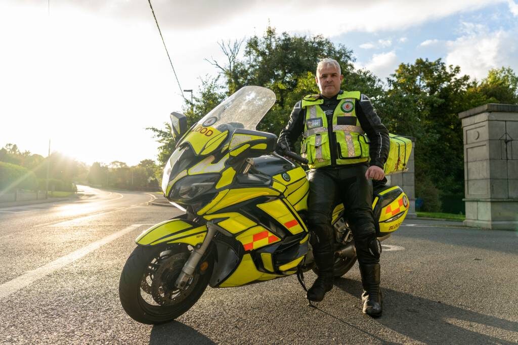 A Blood Bikes volunteer in high-vis beside his motorbike, backlit by low evening sun