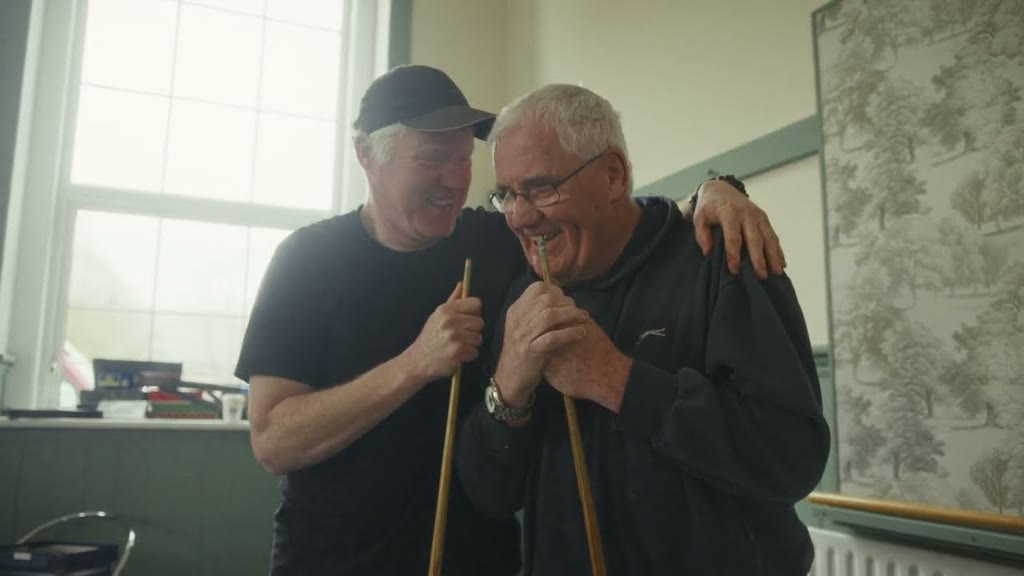 Two older men laughing with their arms around each other, holding snooker cues in a community hall