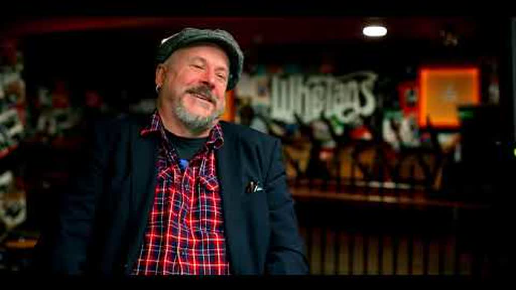 A man in a flat cap and checked shirt sitting smiling inside a music venue