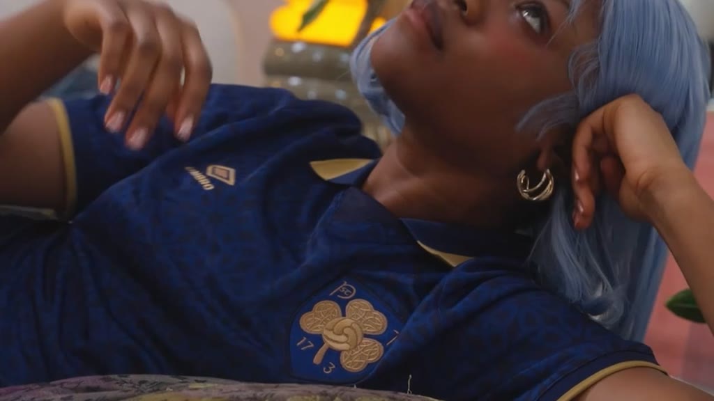 A person with pale blue hair lying back in a navy Umbro shirt with a gold shamrock crest